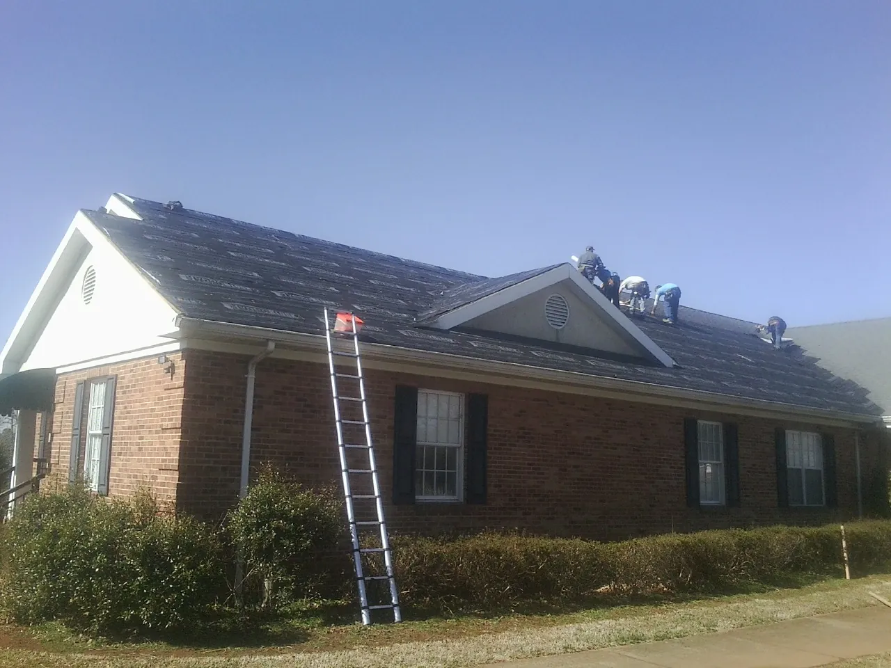 Crew tearing shingles off a brick ranch roof with a ladder set — Emergency Roof Repair work by our crew in Fleming Island