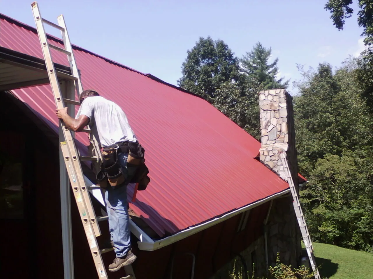 Roofer on a ladder beside a red metal roof and stone chimney — Soffit & Fascia Repair by Roofing Brothers in Fleming Island
